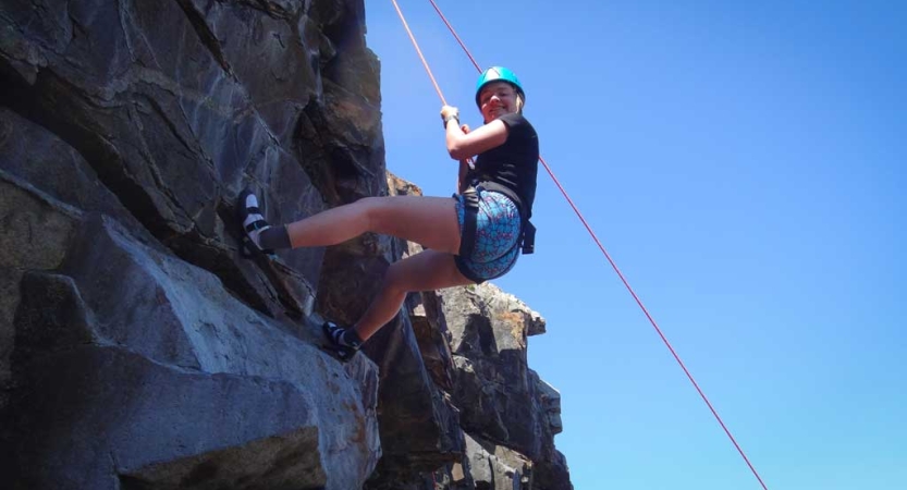 A person wearing safety gear is secured by ropes as they look down at the camera while rock climbing